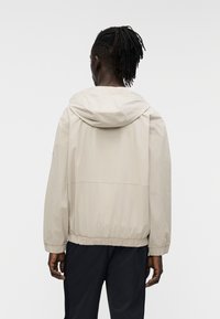 Ανοιχτό μπεζ μπουφάν με κουκούλα, ελαστικές μανσέτες και ζώνη στη μέση. Κατασκευασμένο από λείο ύφασμα, με απλό σχέδιο χωρίς μοτίβα ή διακοσμητικά.