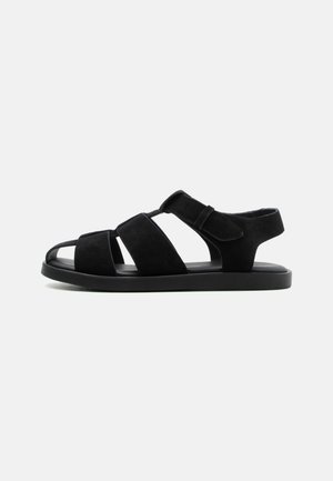 Black flat sandal with three wide straps and a Velcro ankle strap, open toe, and cushioned sole on a plain light background.