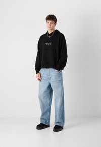 Schwarzer Kapuzenpullover mit einer frontalen Tasche, ausgestattet mit einem zentralen Grafikdesign. Kombiniert mit hellblauen, weiten Jeans und schwarzen klobigen Schuhen.