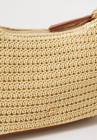 Sac bandoulière tissé beige avec une fermeture éclair dorée et un petit logo doré à l'avant sur fond blanc.