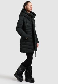 Čierna prešívaná bunda s veľkou kapucňou, horizontálnym prešívaním a bočnými vreckami. Kombinovaná s čiernymi izolačnými snehovými čižmami.