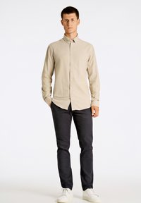 Camisa de botones beige claro con cuello, mangas largas y una tela texturizada, combinada con pantalones oscuros y zapatillas blancas.