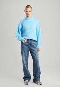 Pull en maille bleu clair avec une finition texturée, des poignets côtelés et un col rond, associé à un jean bleu ample et des baskets.