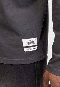 Tricou din bumbac gri cu o margine dreaptă. Prezintă două etichete: "UNBRANDED OUTLAW" și "REDEFINED REBEL", cusute pe partea de jos, în stânga.
