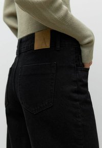 Mustade denim püksid keskmise kõrguse taljega, tagakottide ja pruunist nahast plaastri ning vöökoha peal. Tekstureeritud ja kaunistusteta disain.