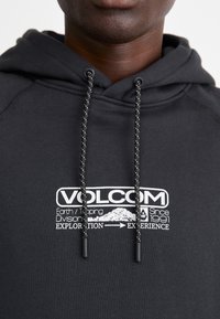Fekete kapucnis pulóver, amelyen egy fehér Volcom logó és felirat látható. Felszerelt egy szürke részletekkel ellátott zsinórral és textúrált anyaggal.