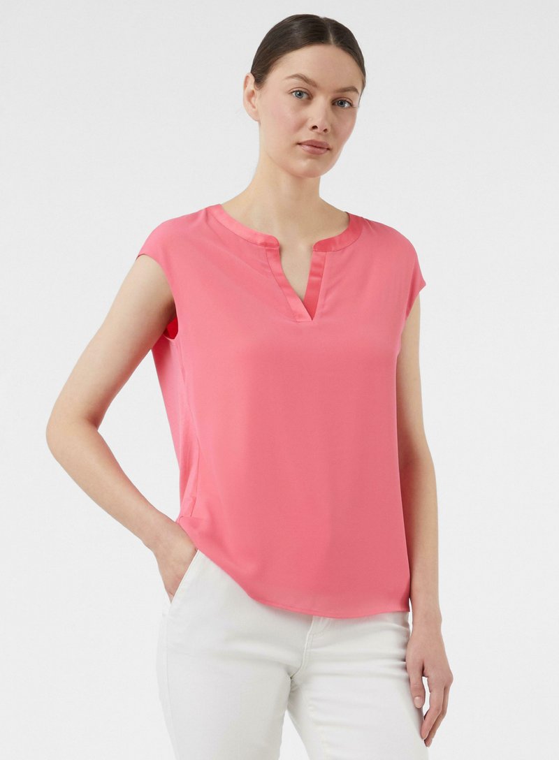 Roze blouse met korte mouwen en een V-hals, gemaakt van een soepele stof. Heeft subtiele zij-spleten en een losse pasvorm. Gecombineerd met witte broeken.