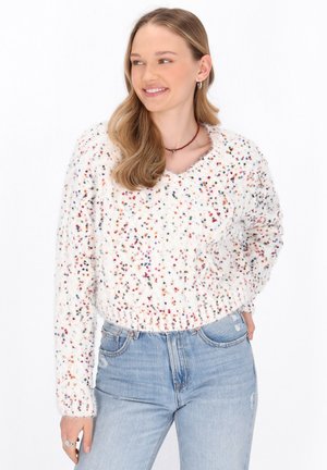 Maglione bianco peloso con macchie multicolori, orlo e polsini a coste; abbinato a jeans azzurri strappati; orecchini rotondi semplici.