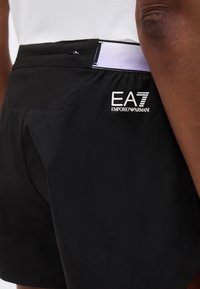 Κοντινό πλάνο ενός ατόμου που φοράει μαύρα σορτς EA7 Emporio Armani με μικρή τσέπη με φερμουάρ και λευκή λεπτομέρεια στη μέση.
