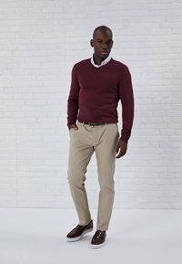 Bărbat stând lângă un zid din cărămidă albă, purtând un pulover vișiniu, cămașă albă cu guler, pantaloni bej și pantofi slip-on maro, având o mână în buzunar.