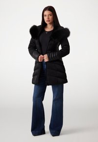 Black puffer coat with a faux fur hood, quilted texture, ribbed sleeves, and a front zipper. Paired with flared dark blue jeans.