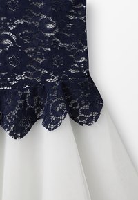 Námořnický krajkový top s květinovým vzorem a vlnkovaným lemem, spárovaný s bílou tylovou sukní. Jemná textura a vrstvené designové prvky.