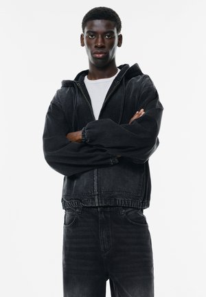 Black denim jacket with hood, featuring elastic cuffs and bottom hem. Worn over a white shirt, paired with black denim pants.