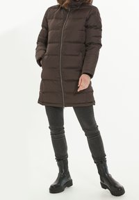 Brązowa kurtka puchowa z zamkiem błyskawicznym, długimi rękawami, o długości do kolan i wysokim kołnierzem, noszona z szarymi dżinsami i czarnymi botkami.