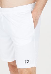 Man med vita elastiska shorts med fickan i högra sidan, visar svart "FZ"-logotyp nära fållen på vänster ben.