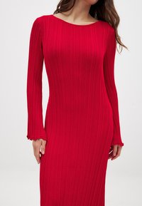 Rød ribbet kjole med lange ærmer, der har en bådformet udskæring og bølgede ærmekanter. Fremstillet af strækbart, tekstureret stof.