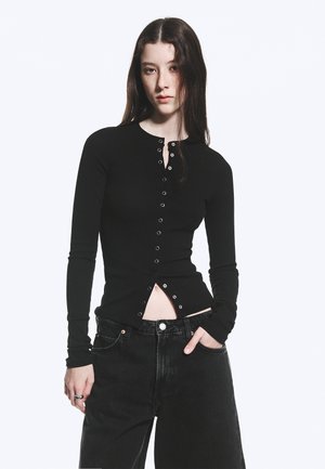 Młoda kobieta z długimi ciemnymi włosami nosi dopasowaną czarną koszulę z długim rękawem i czarne luźne dżinsy, pozując z jedną ręką w kieszeni.