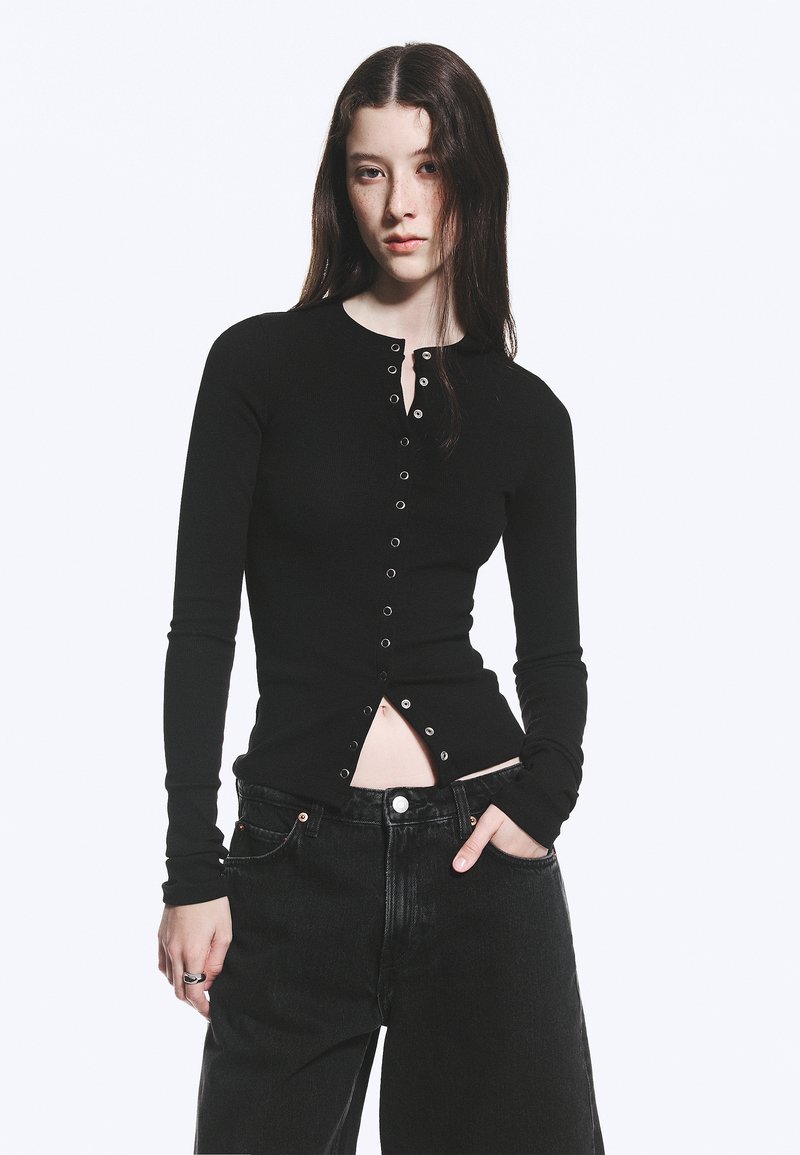 Jeune femme aux longs cheveux foncés porte un haut noir ajusté à manches longues boutonné et un jean noir ample, posant avec une main dans la poche.
