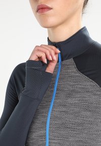 Szary i ciemnoniebieski stanik sportowy zapinany na zamek, o teksturowanym wykończeniu, z niebieskim zamkiem i mankietami z otworami na kciuki. Zbliżenie na obszar kołnierza.