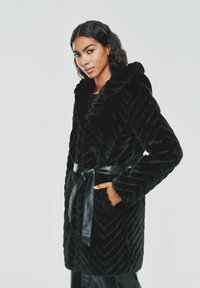 Czarny płaszcz z sztucznego futra w wzór chevron, z kapturem i długimi rękawami. Posiada dodatkowo związywany skórzany pasek oraz dwie boczne kieszenie.