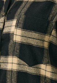 Koszula w czarno-kremową kratę wykonana z miękkiego materiału, z zapięciem na guziki z przodu i kieszenią na piersi, podkreślająca teksturowany, tkany wzór.