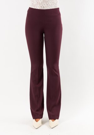 Donna che indossa pantaloni viola scuro a vita alta, aderenti con una leggera svasatura in fondo e scarpe bianche con punta appuntita.