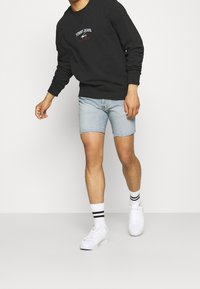 Muž v černé mikině Tommy Jeans, světlých modrých denimových šortkách, bílých ponožkách s černými pruhy a bílých teniskách, jde vpřed.