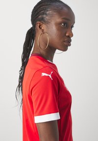 Sarkans sporta krekls, izgatavots no teksturētas auduma, ar baltām akcentēm pie piedurknēm un mazu baltu logo uz pleca.