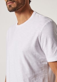 Vit crew-neck t-shirt i lätt, texturerad tyg, med korta ärmar och en avslappnad passform, som visar subtila färgvariationer.