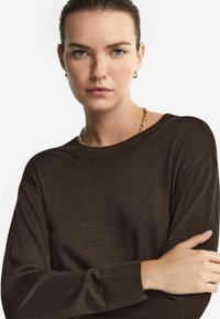 Dunkelbrauner Strickpullover mit rundem Halsausschnitt, gerippten Bündchen und glatter Textur, getragen von einer Person mit kurzem Haar und dezentem Make-up.