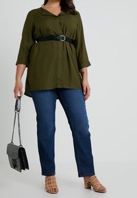 Tunic verde oliva con chiusura a bottoni e cintura, abbinata a jeans dritti blu scuro. Il modello tiene in mano una borsa nera con trama.