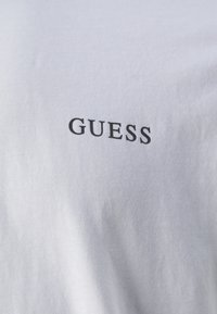 Valge puuvillane T-särk, mille ees on must "GUESS" logo, sujuva tekstuuriga ja tavalise ümmarguse kaelusega disain.