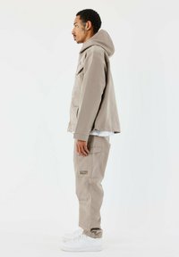 Chaqueta beige con capucha, bolsillos laterales y botones a presión, combinada con pantalones cargo a juego y zapatillas blancas, mostrada de perfil.