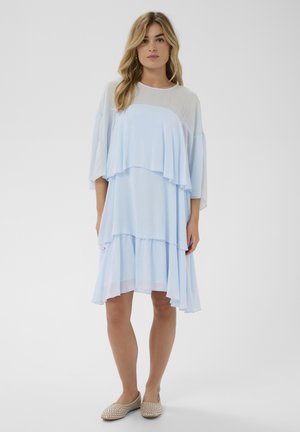 Kobieta w jasnoniebieskiej, warstwowej sukience do kolan oraz beżowych balerinach z ćwiekami, stojąca na tle jednolitego białego tła.