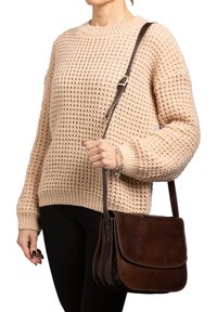 Borsa a tracolla in pelle marrone con patta pieghevole, dettagli di cucitura sottili e una tracolla marrone regolabile, indossata sopra un maglione di colore beige in maglia.