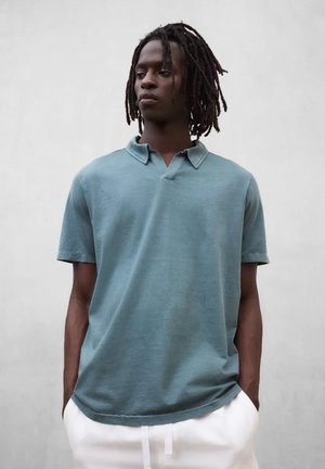 Giovane uomo con dreadlocks fino alle spalle, indossa una polo a maniche corte color teal e pantaloni bianchi, con le mani nelle tasche, guarda verso sinistra.