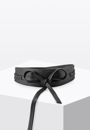 Zwarte leren tailleriem met een gestrikte strik, tentoongesteld op een wit vierkant voetstuk tegen een effen achtergrond.