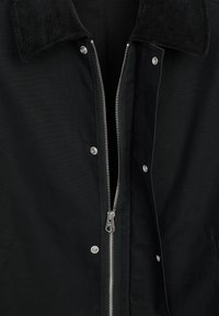 Veste noire avec fermeture éclair argentée partiellement ouverte, boutons-pression devant et un col en velours côtelé visible en haut.