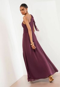 Pikk, varrukateta burgundia värvi kleit külgmise volangiga ja kogutud rinnakuga, millel on voolav seelik ja pehme kangatekstuur, kombineeritud kuldsete kontsadega.