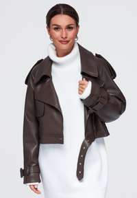 Femme aux cheveux foncés portant des boucles d'oreilles créoles dorées, une robe-pull à col roulé blanche et une veste en cuir marron foncé.