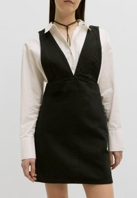 Zwarte denim jurk met een diepe V-halslijn, aansluitend lijfje en een uitlopende rok. Gestyled over een witte blouse met kraag.