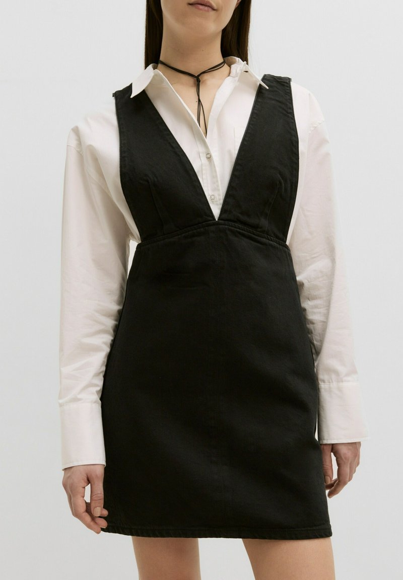 Zwarte denim jurk met een diepe V-halslijn, aansluitend lijfje en een uitlopende rok. Gestyled over een witte blouse met kraag.