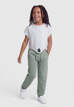 Uśmiechnięte dziecko z zaplecionymi warkoczami nosi białą przyciętą koszulkę z wiązaniem z przodu, zielone spodnie dresowe i białe buty wsuwane, idąc do przodu.