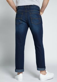 Vaqueros de denim azul oscuro con pierna ajustada, puños enrollados y dos bolsillos traseros con costuras contrastantes. Combinados con zapatillas blancas.