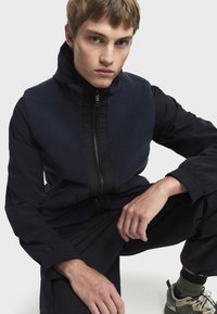 Mornarska fleece prsluk s crnim detaljima od tkanine, zatvaranje na zip, i džepovima; model nosi tamne hlače i robuste tenisice u prigušenim tonovima.