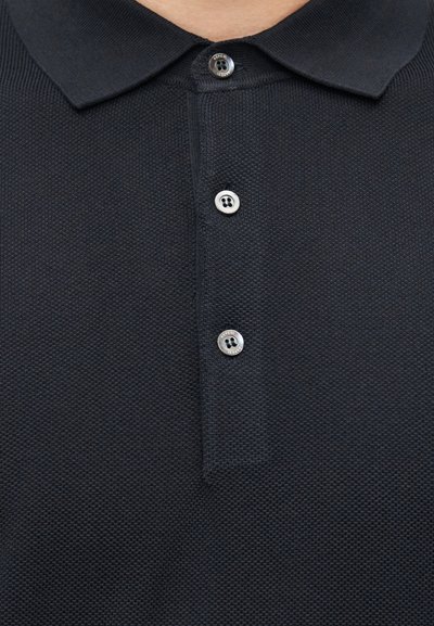 Gros plan sur un polo noir texturé avec trois boutons gris et un col plié.