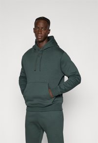 Hoodie verde scuro realizzato in morbido tessuto, con tasca frontale, cappuccio regolabile e piccolo logo del marchio sul petto. Taglio aderente.