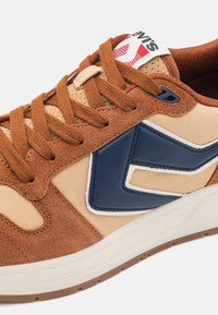 Baskets marron et beige avec logo en cuir bleu, lacets marron, semelle blanche rembourrée et étiquette de languette de marque partiellement visible.