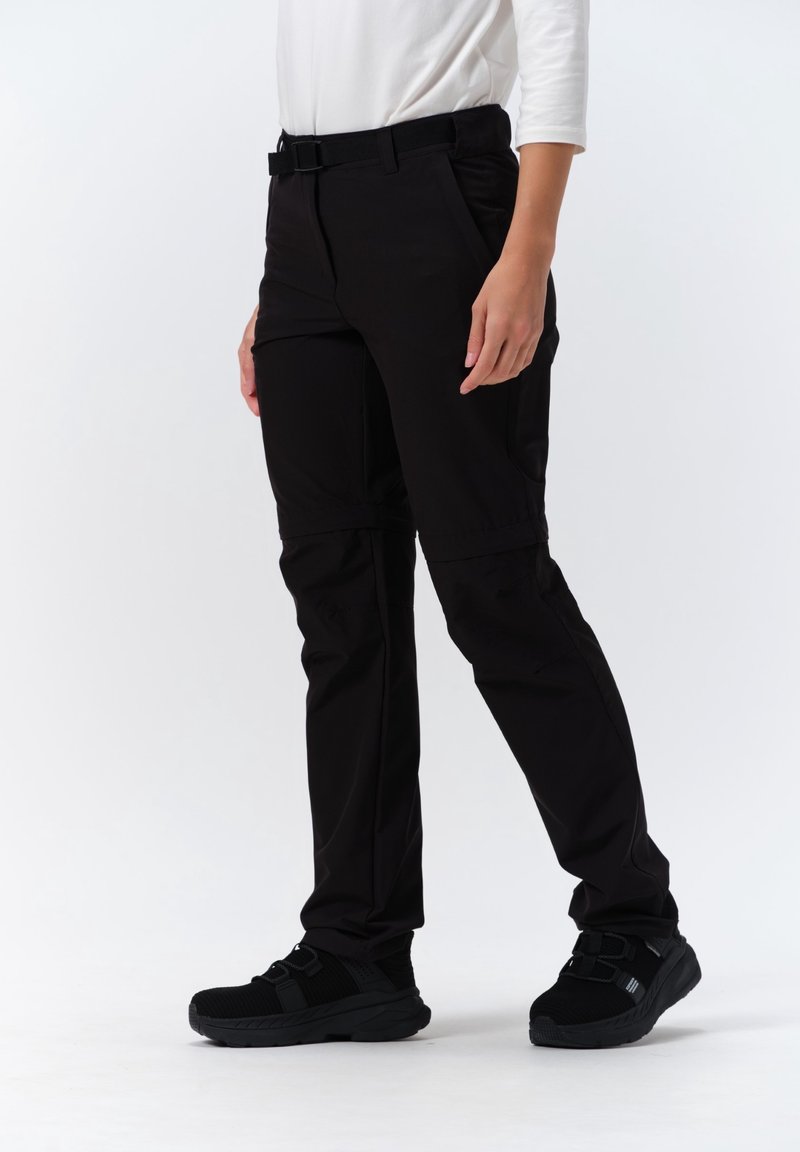 Personne portant un pantalon de randonnée noir avec fermeture éclair, des baskets noires et une chemise blanche à manches longues, debout sur un fond blanc.