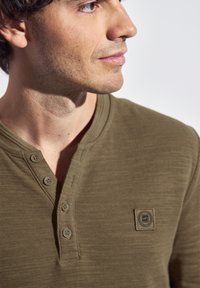 Olivgrön henley-tröja gjord av texturerad tyg, med en tre-knapps placket och en fyrkantig logotyp-patch på bröstet.